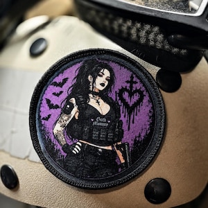 May include: A circular embroidered patch featuring a character with long black hair, wearing a black tactical vest with the text Goth Mommy, set against a purple background with black bats and a dripping heart cross symbol.