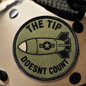 May include: A circular olive green embroidered patch featuring a black graphic of a bomb with a star insignia. The text THE TIP DOESNT COUNT is stitched in black capital letters around the bomb. The patch has a black stitched border.