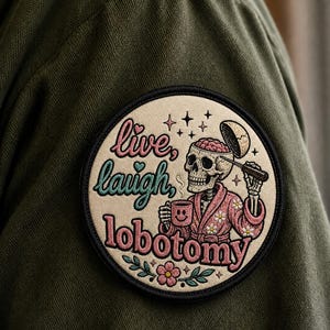 Puede incluir: Un parche bordado circular que muestra un esqueleto con una bata floral rosa sosteniendo una taza de café y una herramienta en su cráneo abierto. El texto dice "live, laugh, lobotomy" en letras rosas y verde azulado sobre tela verde oliva.