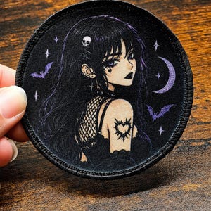 May include: A circular embroidered patch featuring an illustration of a person with long black hair, a skull hair clip, and a heart tattoo on their arm. The background is black with purple bats, a crescent moon, and small white stars.