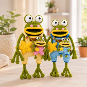 May include: Two large green crocheted frogs with wide mouths and white eyes, each wearing overalls and holding a yellow star. The left frog wears yellow overalls with a small pink-clad frog, while the right wears blue overalls with a small blue-clad frog.