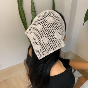 Puede incluir: Un pañuelo triangular de ganchillo en color crema que presenta un patrón de rejilla abierta intercalado con bloques sólidos y cuadrados con textura. El accesorio está hecho de hilo y muestra un diseño geométrico repetitivo.