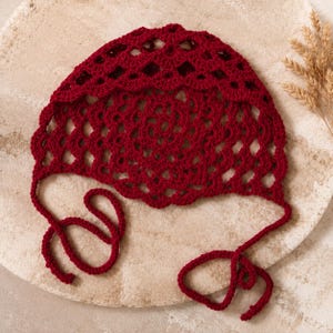 May include: A deep red crochet bonnet featuring an intricate openwork pattern with a scalloped edge and two long matching ties at the base. The headpiece is constructed from yarn with a visible knotted texture and a consistent dark crimson hue.