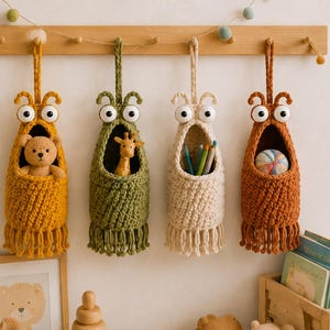 May include: Four hanging crochet storage baskets in mustard yellow, olive green, cream, and terracotta orange. Each basket features two large white eyes, a wide open mouth for storage, and a fringed bottom edge, suspended from a wooden rack.