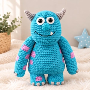Puede incluir: Un muñeco monstruo de ganchillo con cuerpo azul brillante y parches rosas dispersos, dos cuernos grises, grandes ojos de plástico blanco y negro, una amplia sonrisa cosida y un solo diente blanco. La criatura tiene garras grises en manos y pies.