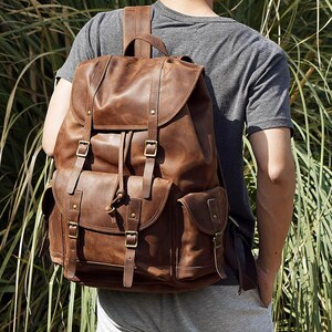 May include: A brown leather backpack featuring a large main compartment with a flap closure, a drawstring top, and two vertical straps with brass-toned buckles. It includes a front pocket and two side pockets, all secured with buckled leather straps.