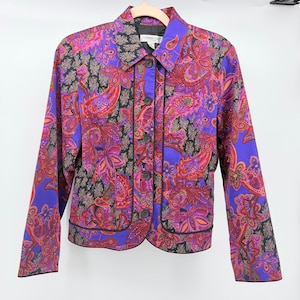 May include: A long-sleeve button-up jacket featuring a vibrant paisley pattern in shades of purple, pink, orange, and black. The garment includes a pointed collar, a front button closure, and black piping trim along the placket and hemline.