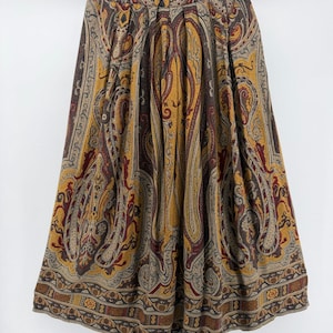 May include: A midi-length skirt featuring a prominent paisley pattern in shades of mustard yellow, deep burgundy, and dark brown. The skirt has a high-waisted design with vertical pleats that create a full, flared silhouette and a wide decorative border.