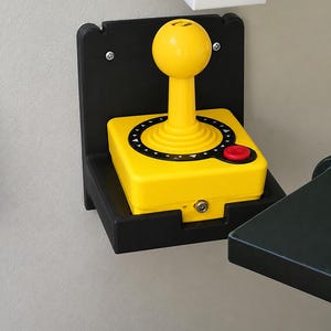 Könnte beinhalten: Ein gelber Arcade-Joystick-Controller mit rundem Kopf, einer schwarzen kreisförmigen Basisplatte mit weißen Dreiecksmarkierungen und einem einzelnen roten Knopf. Der Controller ist in einer schwarzen Kunststoff-Wandhalterung mit zwei silbernen Schrauben befestigt.