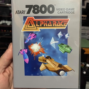 Puede incluir: Una caja de cartucho de videojuego Atari 7800 para el juego Alpha Race. La portada presenta una escena de batalla espacial con una nave espacial color canela, una nave verde y una nave rosa sobre un fondo azul oscuro estrellado con una explosión.