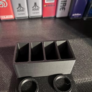 Könnte beinhalten: Ein mattschwarzer, 3D-gedruckter Kunststoff-Organizer mit vier rechteckigen vertikalen Schlitzen in der hinteren Reihe und zwei kreisförmigen Öffnungen in der vorderen Reihe, der kleine Gegenstände oder Spielkassetten aufrecht hält.
