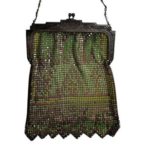 May include: A vintage-style metal mesh handbag featuring a green and dark brown geometric pattern. The bag has a decorative dark metal frame with an ornate floral engraving and a kiss-lock closure, finished with a scalloped bottom edge.