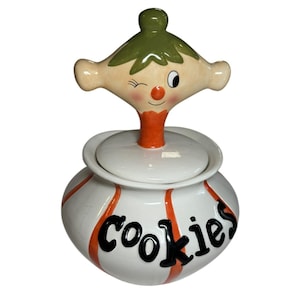 May include: A white ceramic cookie jar with vertical orange stripes and the word Cookies written in black script. The lid features a sculpted figure with a green hat, a winking face, a red nose, and an orange neck, serving as a handle.