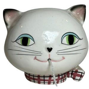 May include: A white ceramic cat head figurine with green eyes, blue eyelids, and a pink mouth. A white string extends from the mouth. The cat wears a red and black plaid collar. The ears have black tips and pink inner details on a glossy finish.