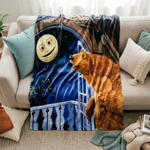 May include: A soft fleece throw blanket featuring a graphic print of a brown bear standing on a balcony at night. The scene includes a large, smiling moon in a dark blue starry sky, a white balcony railing, and a wooden door frame structure.