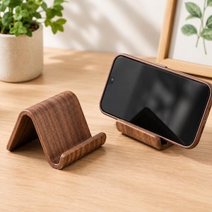Puede incluir: Un soporte para teléfono de madera de color marrón oscuro con veta visible y un perfil curvo en forma de onda. Un soporte está vacío, mientras que el otro sostiene un teléfono inteligente negro en posición horizontal. Tiene un acabado pulido.