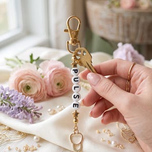 May include: A gold-toned keychain featuring a lobster clasp, a metal key, and a vertical string of white beads spelling the word PULSE. The beads are accented with small gold stars and spheres, and the bottom charm is a gold-toned medical stethoscope.