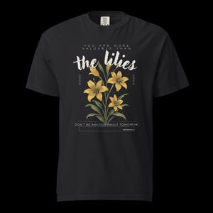 Könnte beinhalten: Ein schwarzes Kurzarm-T-Shirt mit einer Grafik aus gelben Lilien und grünen Blättern. Der Text lautet: YOU ARE MORE VALUABLE THAN the lilies, DON'T BE ANXIOUS ABOUT TOMORROW und MATTHEW 6:28-34. Das Design ist mittig auf der Brust platziert.