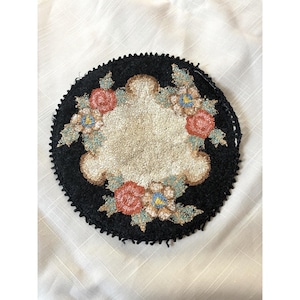 Pode incluir: Um porta-copos circular de punch needle com fundo preto e uma área central texturizada na cor creme. O design é acentuado por motivos florais em tons de coral, azul claro e verde sálvia, com uma borda escura de franjas.