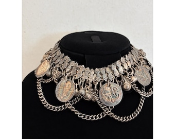 Puede incluir: Un collar tipo gargantilla de tono plateado con una banda ancha y texturizada, adornada con múltiples dijes circulares similares a monedas y pequeñas cuentas esféricas, unidos por delicadas cadenas plateadas que cuelgan en capas.