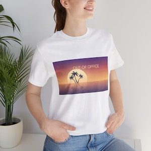 Pode incluir: Uma t-shirt branca de manga curta com uma impressão gráfica retangular de um pôr do sol sobre o oceano com silhuetas de palmeiras e o texto OUT OF OFFICE em letras maiúsculas brancas sobre o sol num céu roxo e cor de laranja.