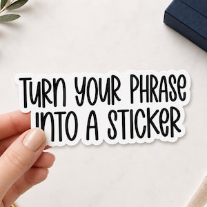 May include: A white die-cut sticker with a black border featuring the text "TURN YOUR PHRASE INTO A STICKER" written in a bold, rounded, sans-serif font. The sticker has a matte finish and is shaped to follow the contour of the lettering.