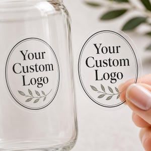 May include: A clear glass bottle featuring a circular transparent sticker with a black double-line border. The sticker displays the text Your Custom Logo in a serif font, accented by a small botanical leaf illustration at the bottom.