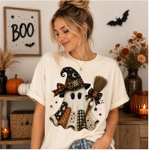 May include: A cream-colored t-shirt featuring a graphic of a ghost wearing a witch hat and holding a broom. The ghost is decorated with various patterns including plaid, polka dots, and floral prints in shades of orange, black, and white.