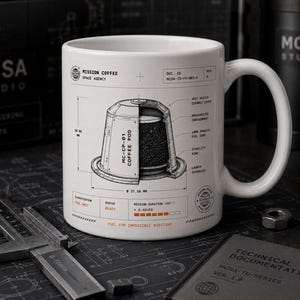 May include: A white ceramic mug featuring a technical diagram of a coffee pod labeled MC-CP-01. The print includes text such as Mission Coffee Space Agency, Fuel for Impossible Missions, and technical specifications with dimensions of 50.84 mm and 37.58 mm.