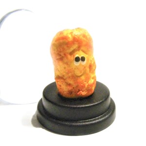 May include: A small, yellow, cartoon-like figurine of a potato with a smiling face and googly eyes. The figurine is standing on a black, circular base.