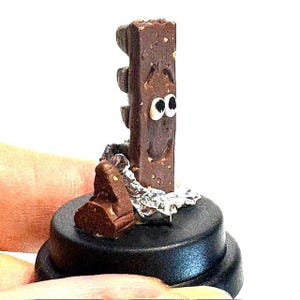 May include: A novelty chocolate bar figure with cartoon eyes and a smiling expression, mounted on a black base. The chocolate is partly wrapped in foil, and a small piece of chocolate sits on the base. The figure is held by a hand.