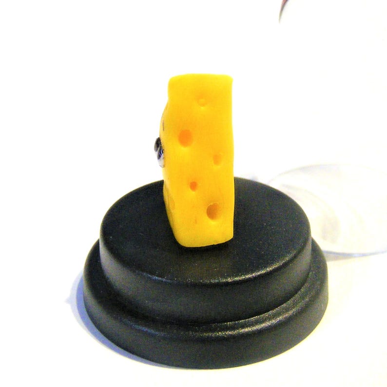 May include: A small, yellow, block of cheese with holes, sitting on a black circular base. The cheese is shaped like a cube and has a smooth surface.