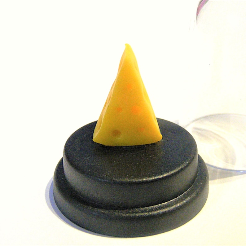 May include: A small, yellow wedge of cheese with holes, displayed on a black pedestal inside a clear glass dome.