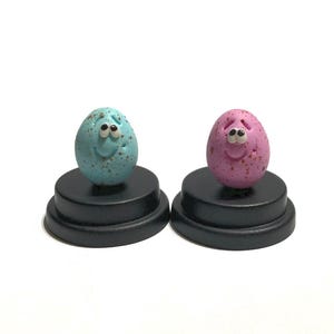 May include: Two small ceramic eggs with painted faces, one blue with brown speckles and one pink with red speckles, are displayed on black bases.