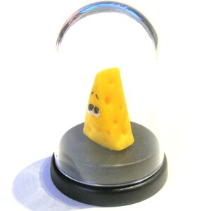 May include: A small, yellow cheese wedge with a cartoon face is displayed in a clear glass dome on a black base.