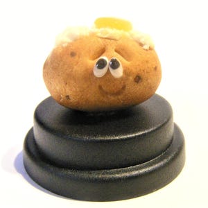 May include: A small, brown potato with a smiling face and googly eyes sits on a black pedestal. The potato has a small piece of yellow butter on top.