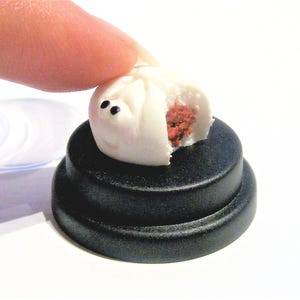 May include: A miniature white and red steamed bun with black eyes, sitting on a black circular display stand.