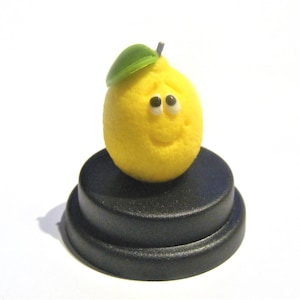 May include: A yellow lemon figurine with a green leaf on top and a smiling face. The figurine is sitting on a black circular base.