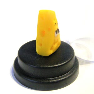 May include: A small, yellow cheese wedge with a cartoon face and holes. The cheese is sitting on a black circular base.