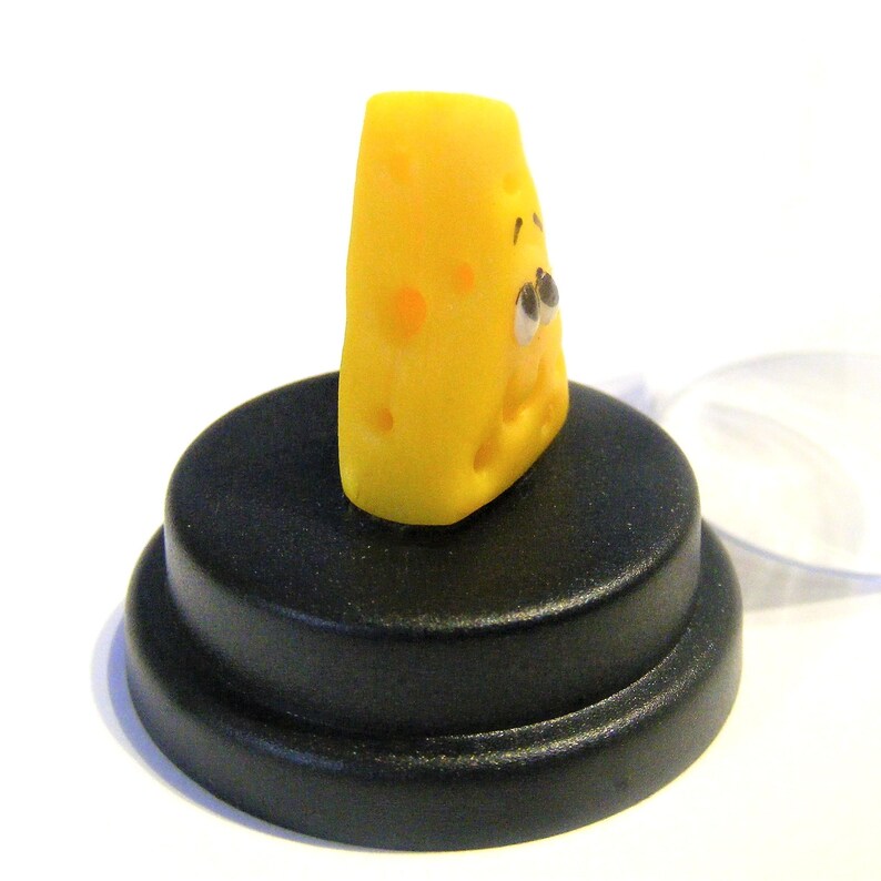 May include: A small, yellow cheese wedge with a cartoon face and holes. The cheese is sitting on a black circular base.