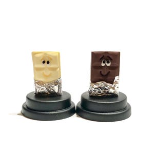 May include: Two miniature chocolate bars, one white and one dark, with smiling faces, are displayed on black bases. The chocolate bars are wrapped in silver foil.