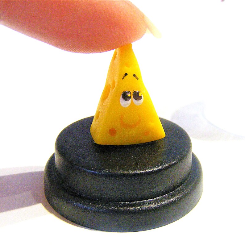 May include: A small yellow cheese wedge with a smiling face and black eyes sits on a black circular base.