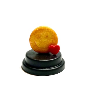Peut inclure: Un biscuit jaune en forme de smiley avec un cœur rouge sur un piédestal noir.