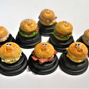 May include: Eight miniature, cartoon-faced,  sandwiches on black pedestals. Each sandwich has a different filling, including ham, cheese, and lettuce.