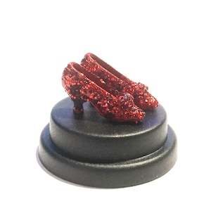 May include: Miniature red glitter shoes with a small heel, displayed on a black, tiered base. The shoes are a bright red with a sparkling texture. The base is a matte black, creating a contrasting backdrop. A decorative item.