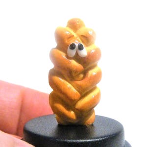 May include: A small, hand-painted ceramic figurine of a braided bread with googly eyes. The figurine is brown and yellow and is standing on a black base.