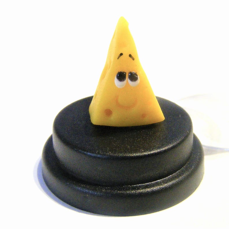 May include: A small yellow cheese wedge with a smiling face and black eyes. The cheese is sitting on a black circular base.