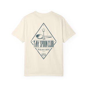 Può includere: Una t-shirt a maniche corte color crema con una grafica blu scuro sul retro. Il design a forma di rombo contiene due cucchiai, il testo "TINY SPOON CLUB" e la frase "THE BIG ONE IS HOSTILE" con un piccolo emblema "CT".