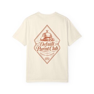 以下が含まれることがあります： クリーム色の半袖Tシャツで、背面に錆色のグラフィックがプリントされています。ひし形のデザインの中には、サービスベル、ベビーソックス、おしゃぶりが描かれており、「Default Parent Club」および「Apparently I Know Everything」というテキストが添えられています。