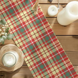 May include: A rectangular table runner featuring a plaid pattern with intersecting lines in shades of red, sage green, pale yellow, and white. The fabric has a woven texture and is displayed flat, showcasing the repeating geometric grid design.